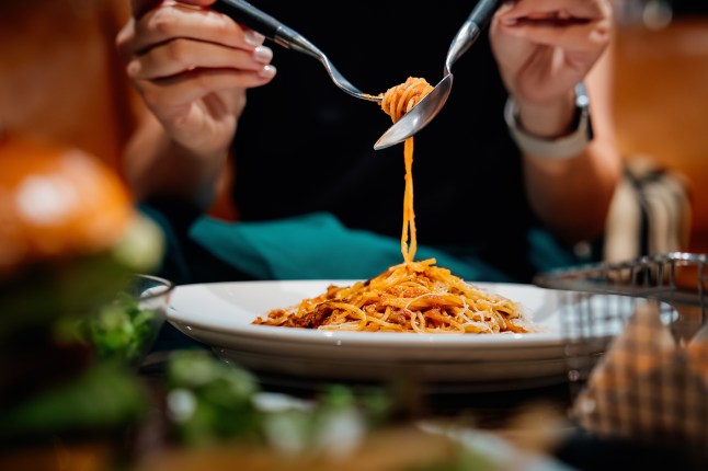 Close-up, mid-section of young Asian woman sitting at dining table holding cutleries, twirling freshly served spaghetti bolognese and enjoying her meal in restaurant. Italian food. Dining out. People, food and lifestyle